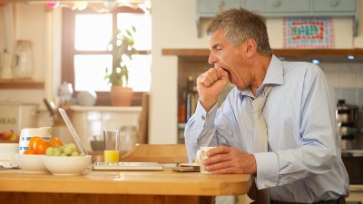 Man yawning whilst sat in kitchen looking at his laptop