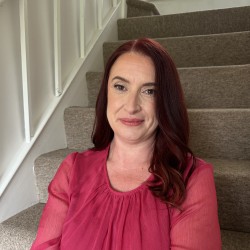 Head and shoulders shot of Charlotte, a white woman in her forties with shoulder-length dark red hair. She is wearing a hot-pink blouse and sitting on a staircase