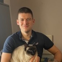 Tom is sitting down on a chair wearing a blue t-shirt, with a cat on his lap.