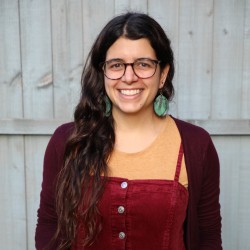 A photo of Dr Violeta Menendez Gonzalez wearing a red corduroy dress, a yellow tshirt, blue large earrings, and brown glasses