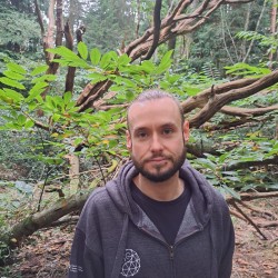 Man with grey hoodie, beard and lip piercing, stood infront of a fallen tree.