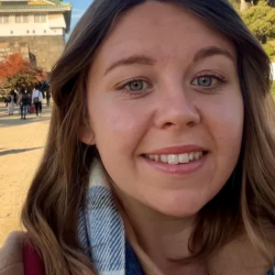 Woman smiling at camera, head and shoulders shot, with sunny buildings and trees in background.