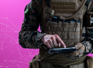 A soldier in combat uniform uses a tablet computer in front of a pink background with network concept lines