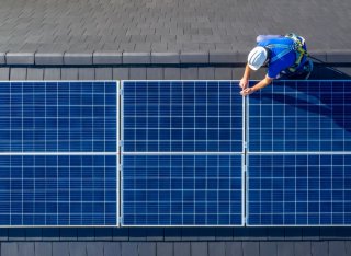 An aerial shot of a person wearing a helmet and blue top working on solar panels on a roof