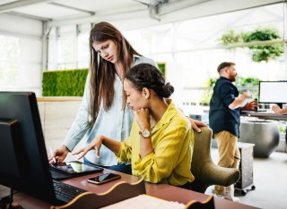 Two female office workers in conversation at a computer.