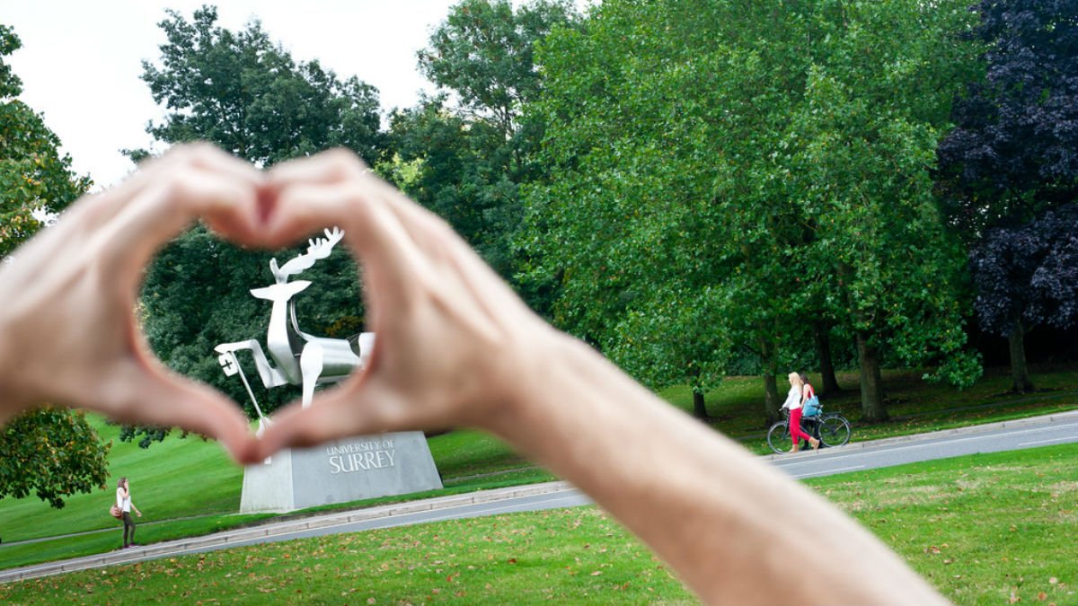 A student makes heart hands around the University of Surrey's stag statue on campus