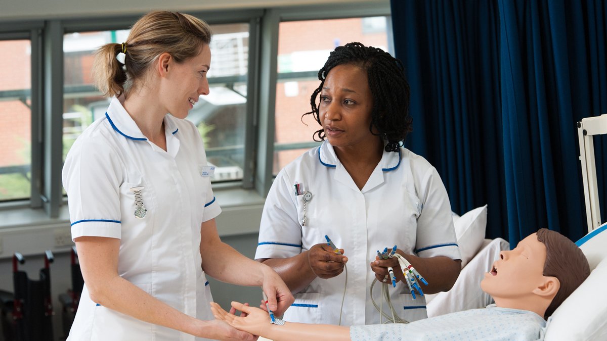 Two nursing students standing next to a mannequin laying on a bed