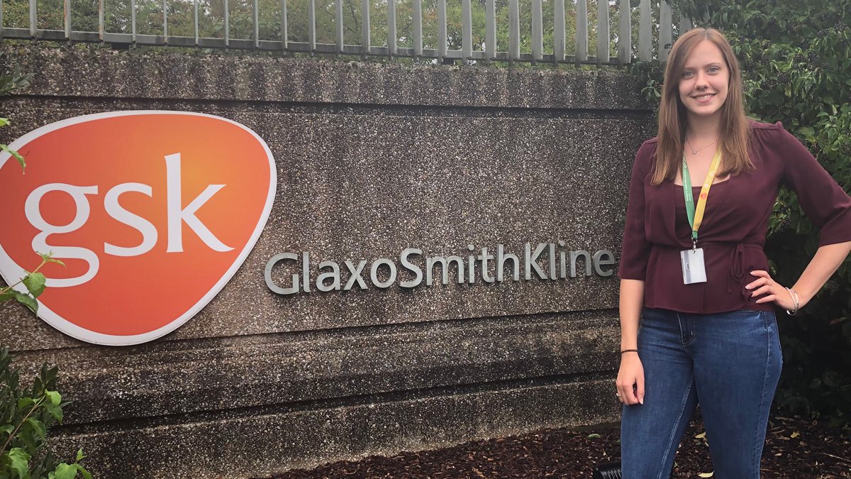 Biosciences student, Chloe Charlwood, standing in front of a GlaxoSmithKline (GSK) sign
