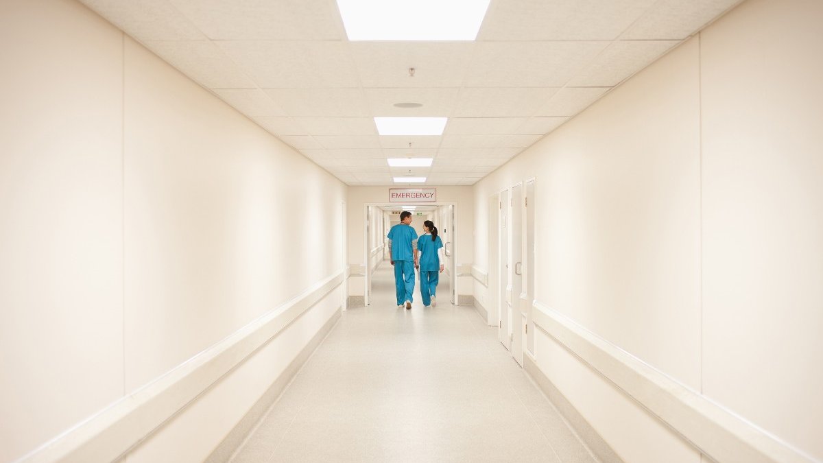 Two people wearing green scrubs can be seen a long way down a bright, white hospital corridor