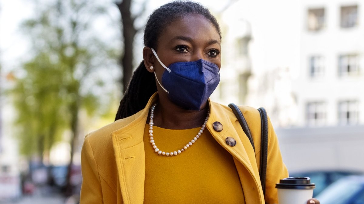 A commuting woman wearing a navy FFP2 face mask, yellow suit and pearl necklace carries a coffee along an urban street