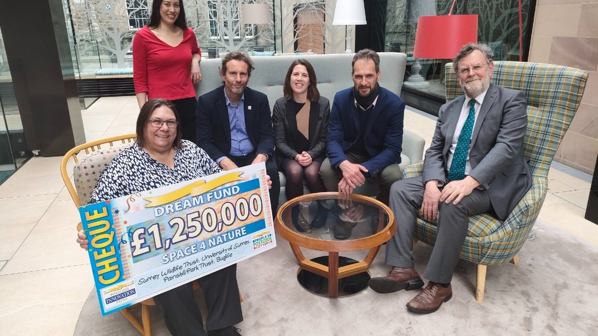 Five people sit on armchairs or a sofa and one stands in a spacious lobby. One of the seated people holds a giant Dream Fund cheque made out for £1,250,000 to Space 4 Nature