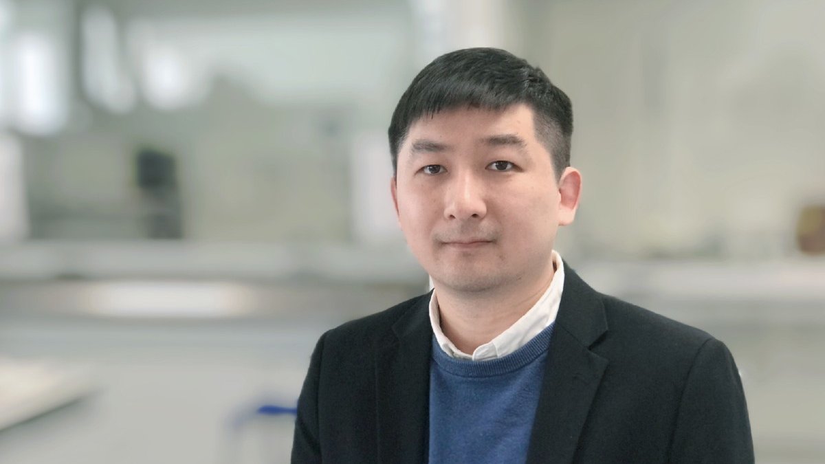 An Asian man wearing a dark jacket, blue jumper and white collared shirt stands in a light, airy building