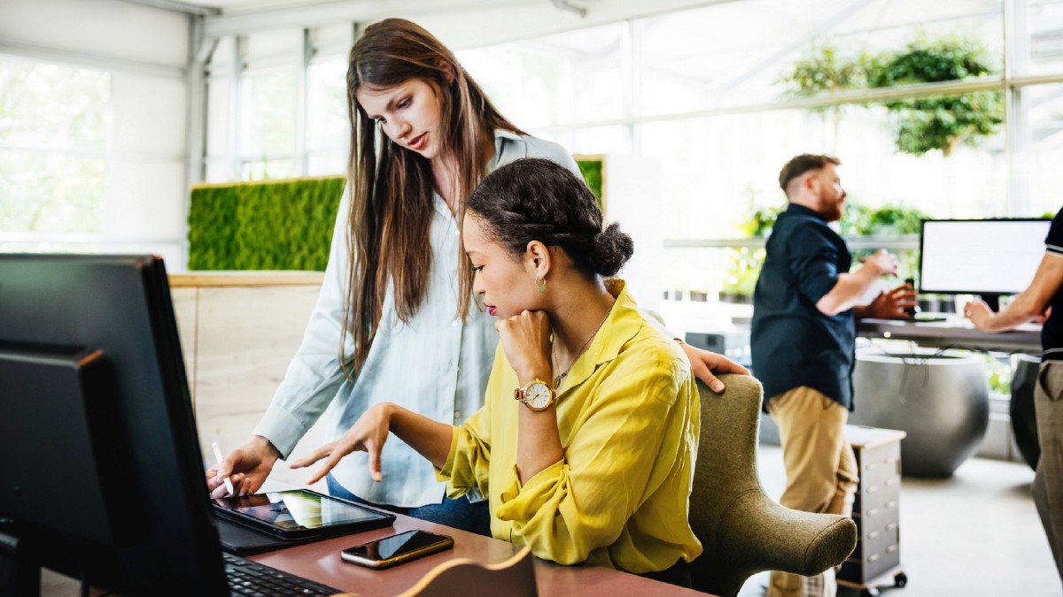 Two female office workers in conversation at a computer.