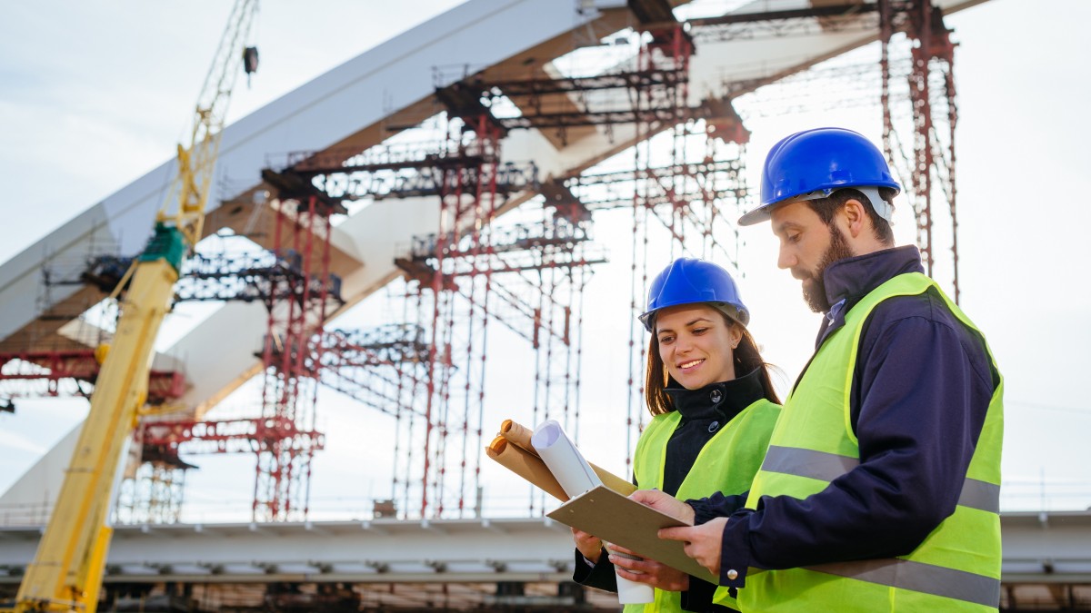 Two engineers, male and female, in protective helmets and reflective clothing, standing on construction site where large arch bridge is being built.