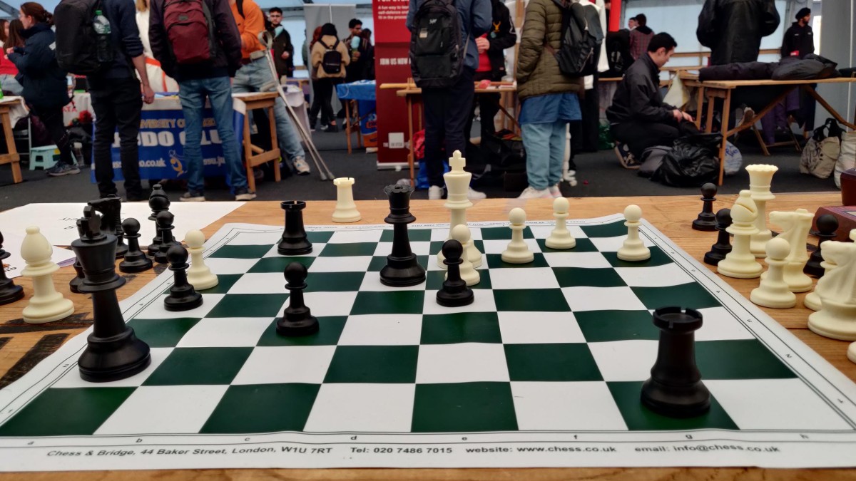 The photo shows a chessboard on a table with students in the background.