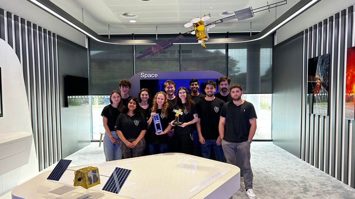 11 students wearing jeans and black t-shirts stand in an office with space paraphernalia around them. Two hold trophies 