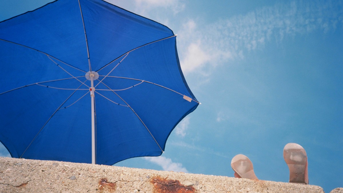 A person sunbathing under a blue parasol with a blue sky.